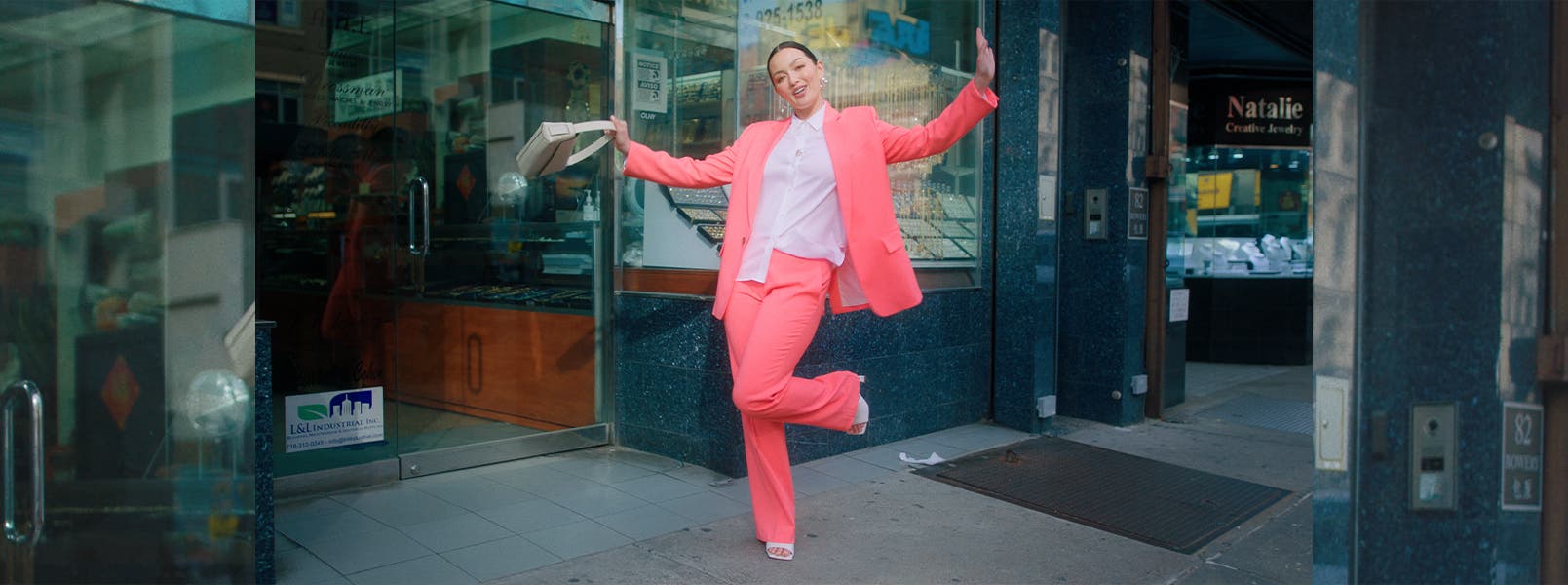 Model Mia Kang wearing a pink pantsuit and white button-up shirt with a cream-colored handbag and white heels.