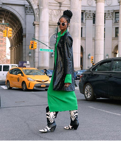 A woman wearing a green dress, black coat and cowboy boots.