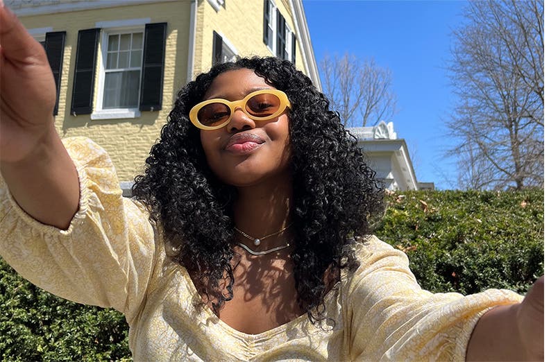 A model taking a selfie outdoors while wearing yellow sunglasses.