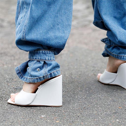 A closeup of a woman wearing white wedge heels with blue jeans.