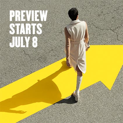 Anniversary Sale preview starts July 8. A woman in platform boots and a cream outer layer walks across a yellow arrow on the street.