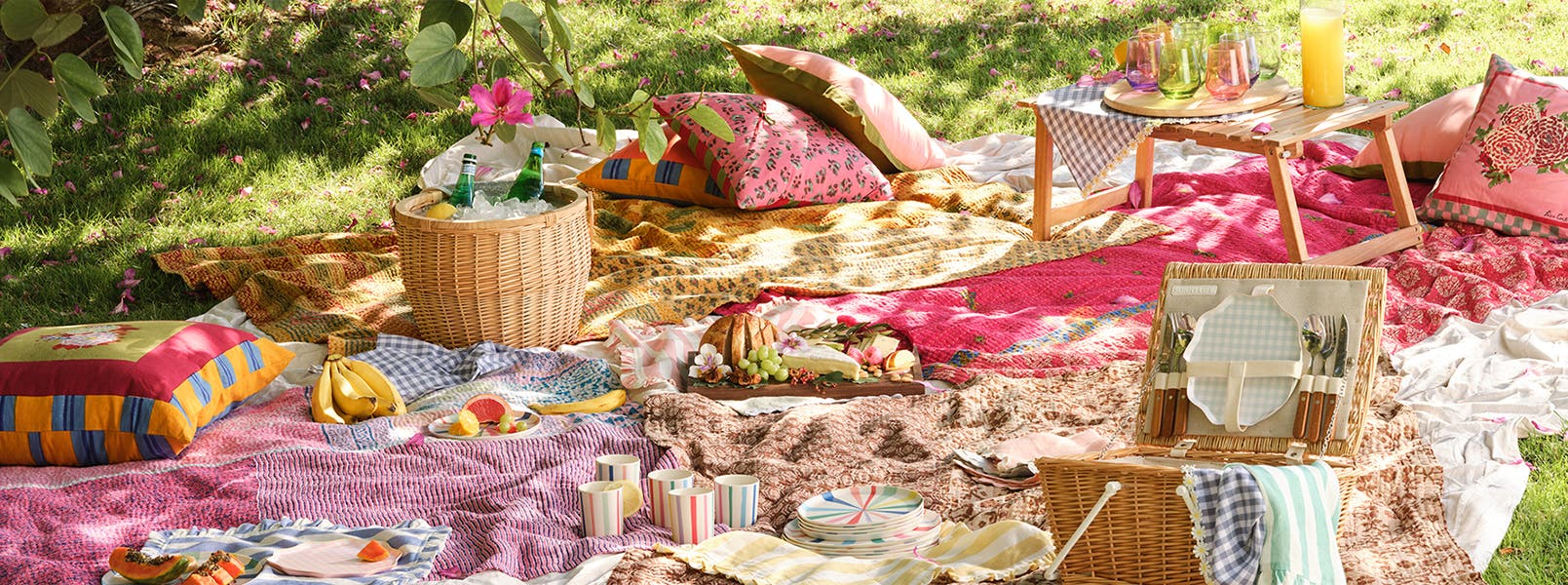 A picnic in the grass with colorful blankets, tablecloths, pillows, tableware and more.
