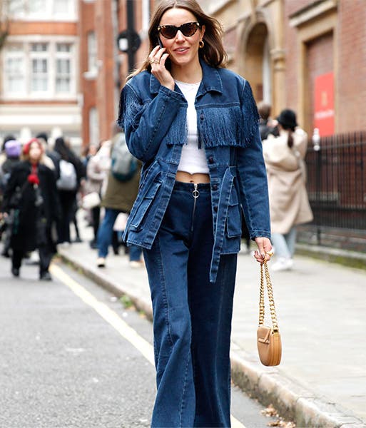 A woman wearing a white cropped tee with a fringed denim jacket and denim pants.