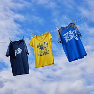 Three college sports team T-shirts hanging on a clothesline with a blue-sky background.