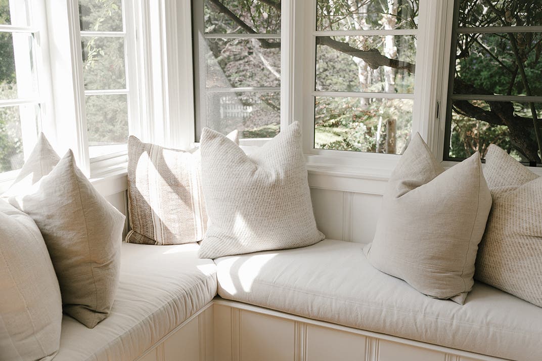 Corner of a windowed room, neutral window seat and pillows. 