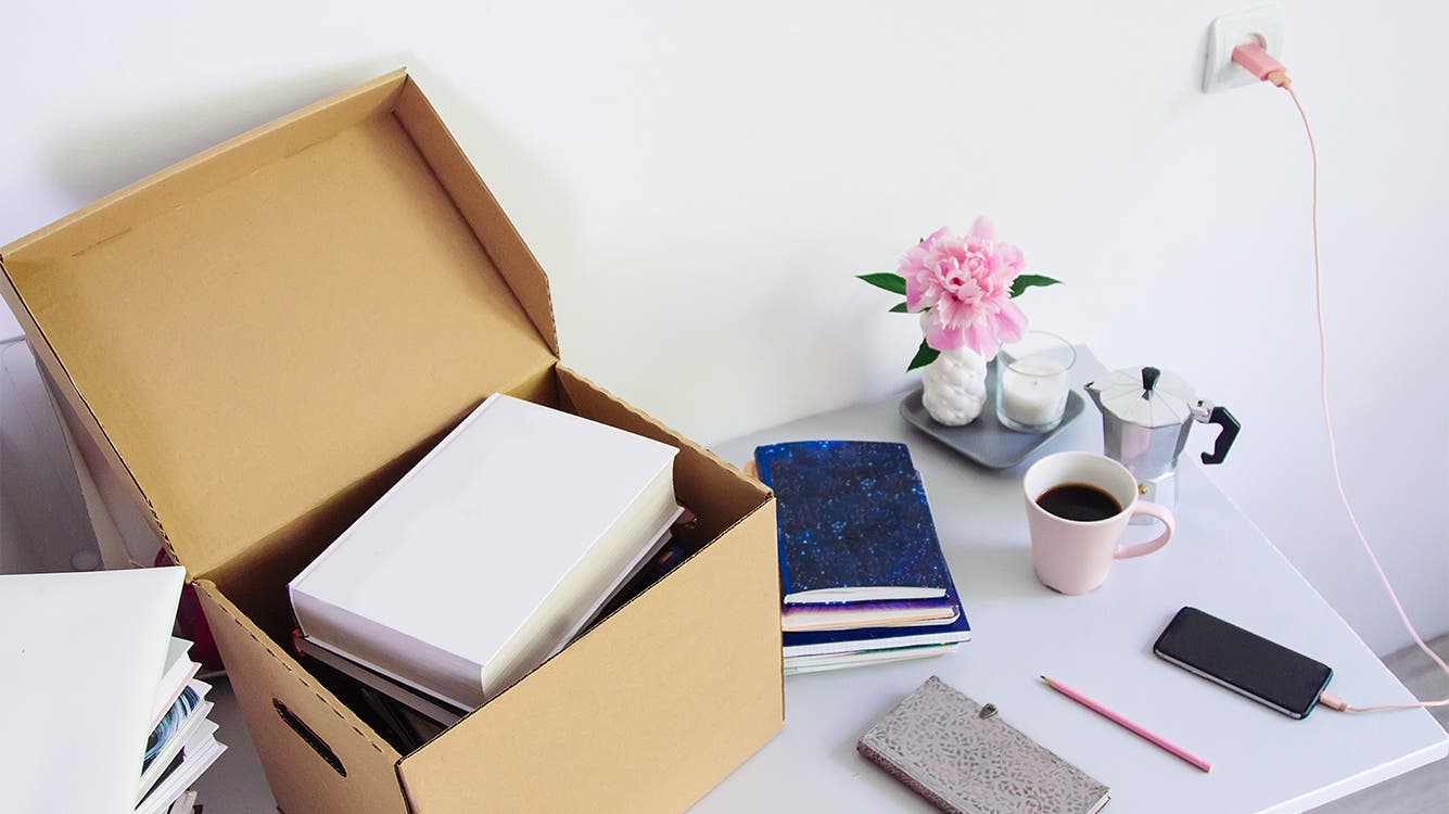 A box of books and a stack of notebooks sit next to a coffee cup and a phone on a desk.