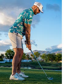 Man playing golf in colorful patterned shirt, khaki shorts, Nike golf shoes and white baseball cap.