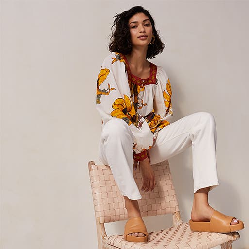 A woman wearing a printed blouse with white jeans and tan sandals.