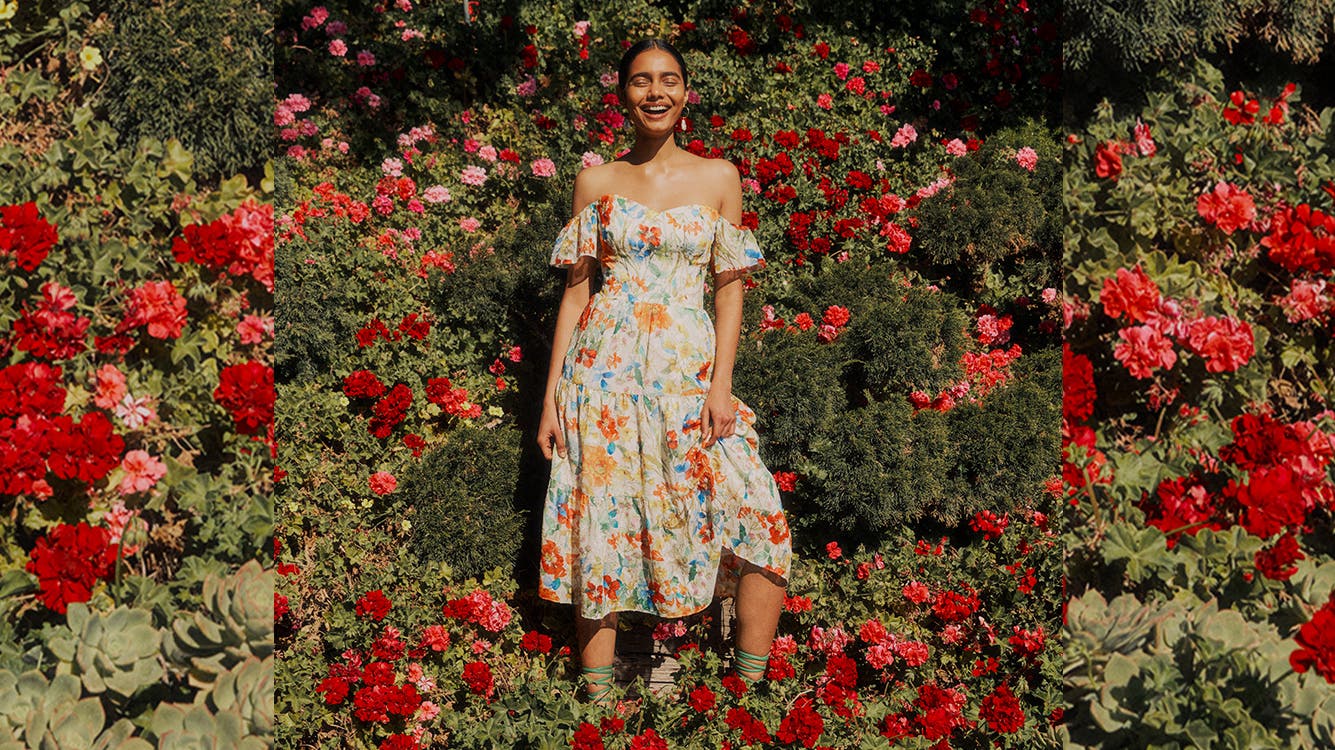 A woman wearing an off the shoulder dress standing in front of lush rose bushes.