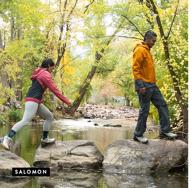 A woman and man wearing outdoor activewear and crossing a stream.