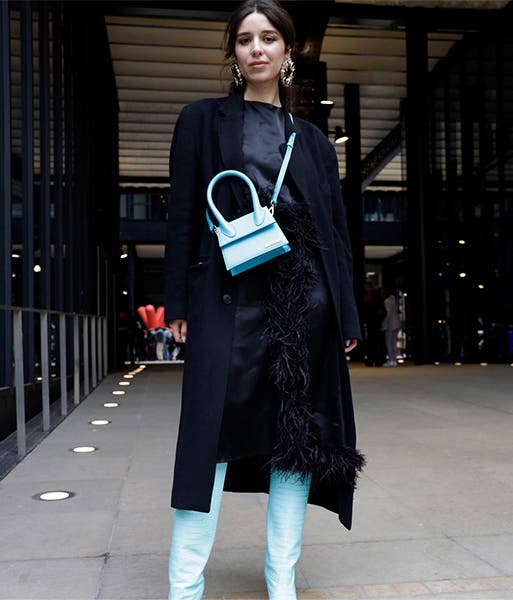 A woman wearing sky blue knee-high boots and a matching crossbody bag with a long black coat and dark top and pants.