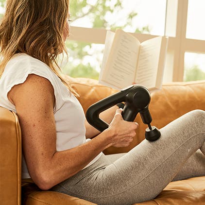 A seated woman using a Theragun percussive therapy massager on her leg.