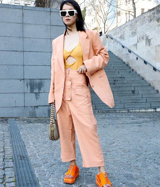 A woman wearing a matching orange creamsicle-colored pant and blazer set with an orange cutout top underneath and bright orange sandals.