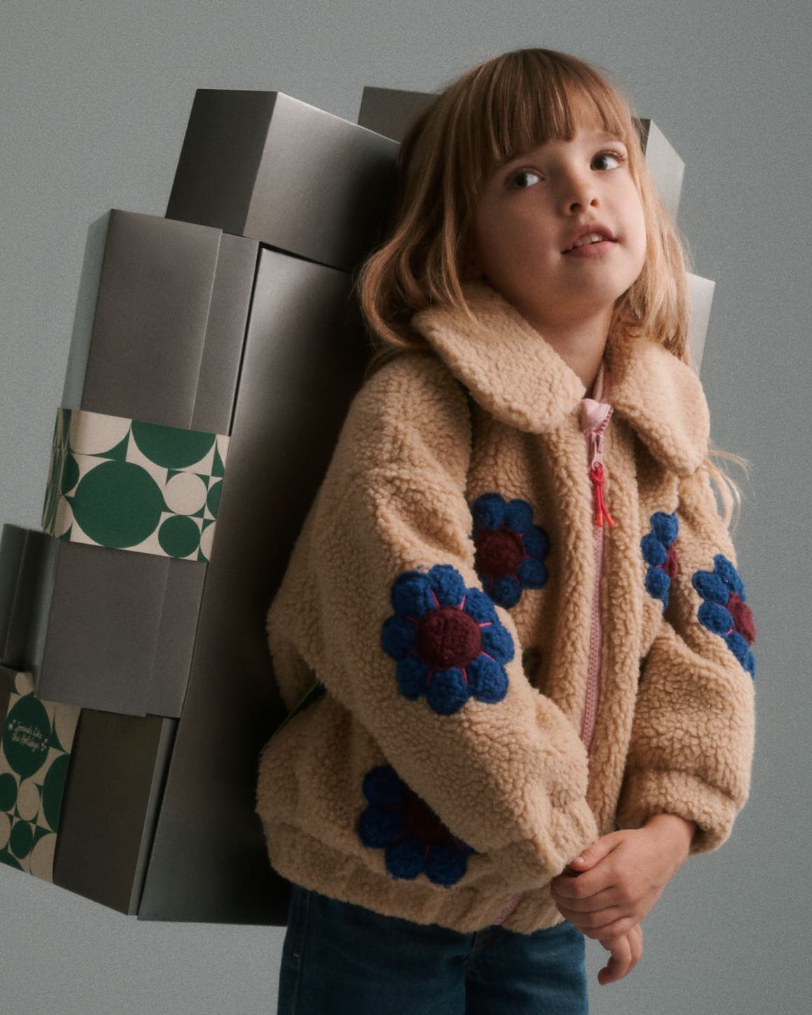 A girl in a fleece jacket carrying a stack of silver Nordstrom gift boxes on her back.