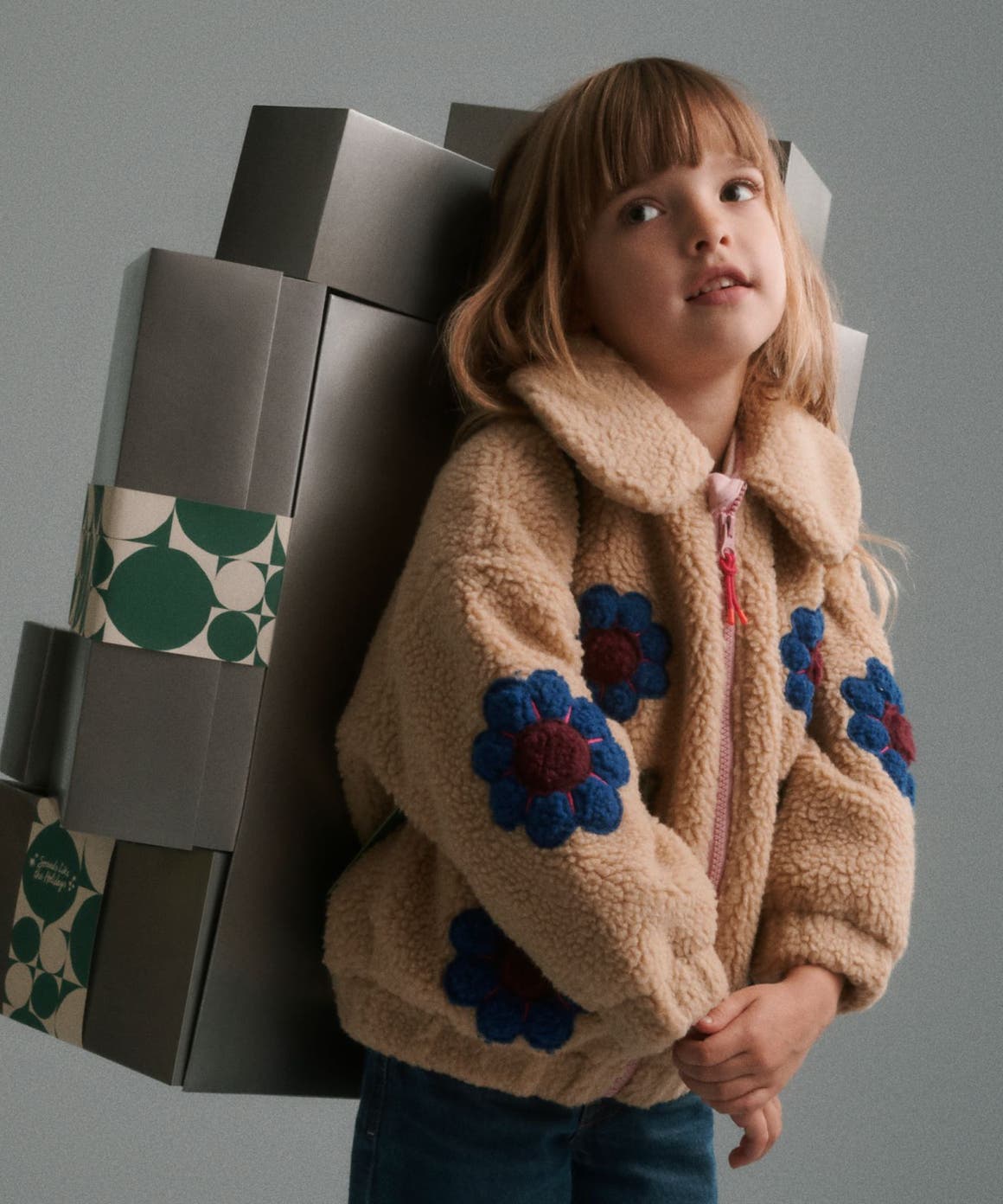 A girl in a fleece jacket carrying a stack of silver Nordstrom gift boxes on her back.