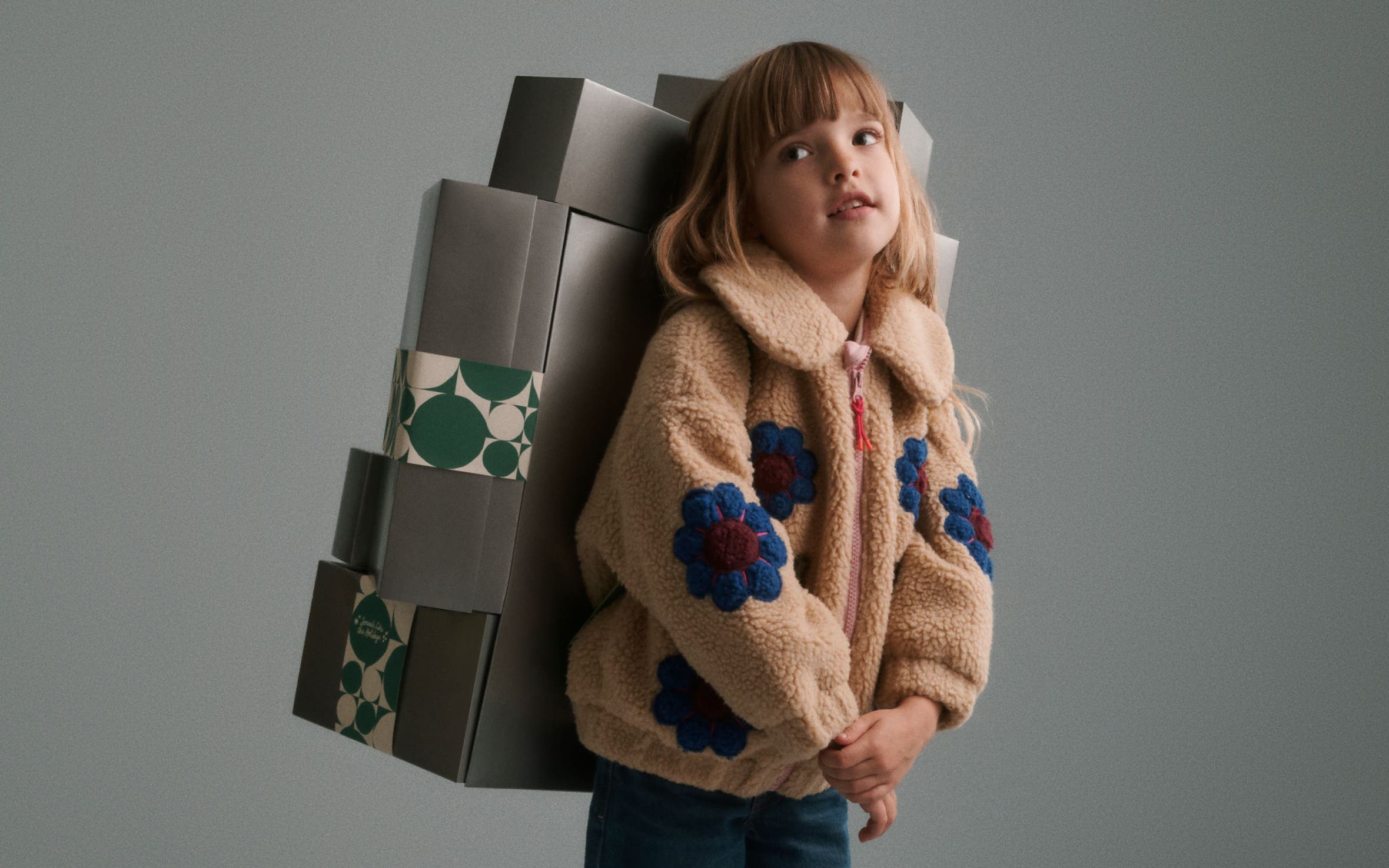 A girl in a fleece jacket carrying a stack of silver Nordstrom gift boxes on her back.
