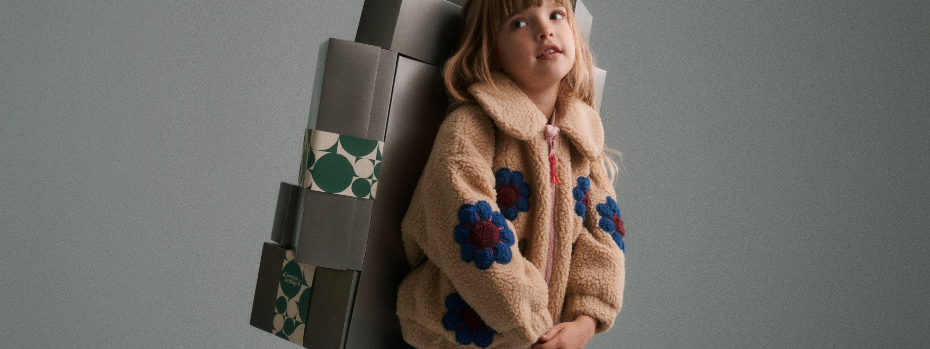 A girl in a fleece jacket carrying a stack of silver Nordstrom gift boxes on her back.