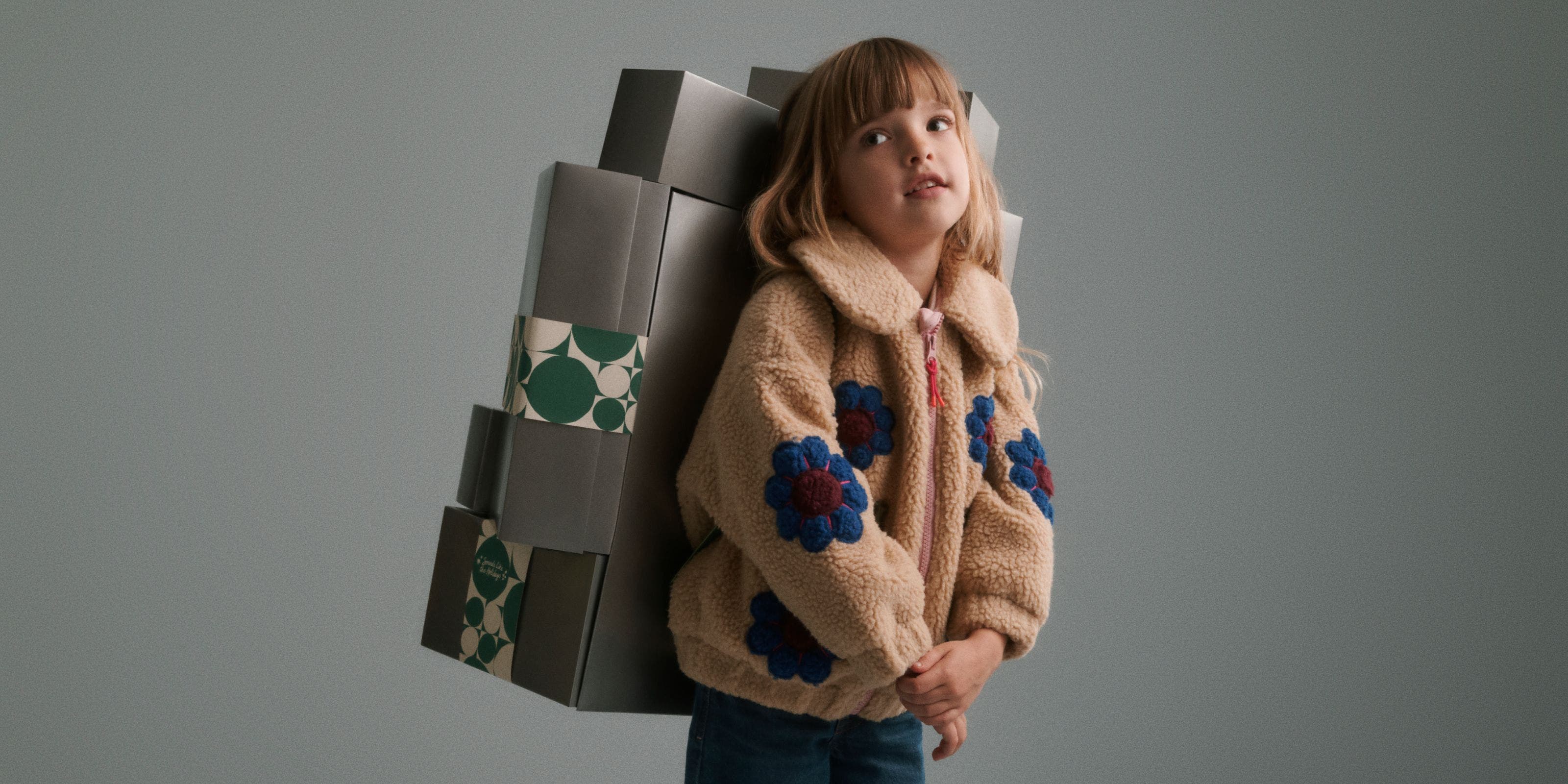 A girl in a fleece jacket carrying a stack of silver Nordstrom gift boxes on her back.