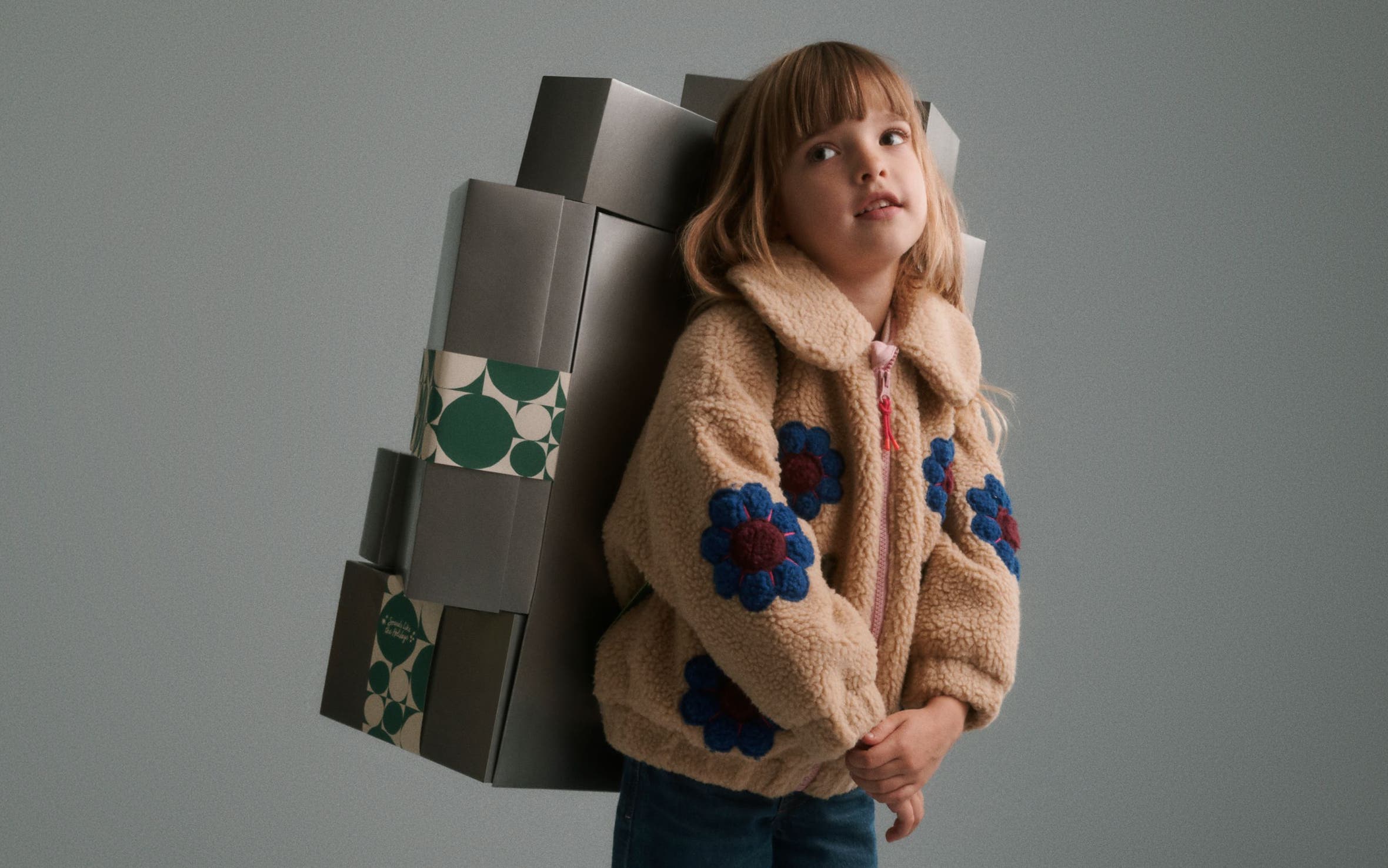 A girl in a fleece jacket carrying a stack of silver Nordstrom gift boxes on her back.