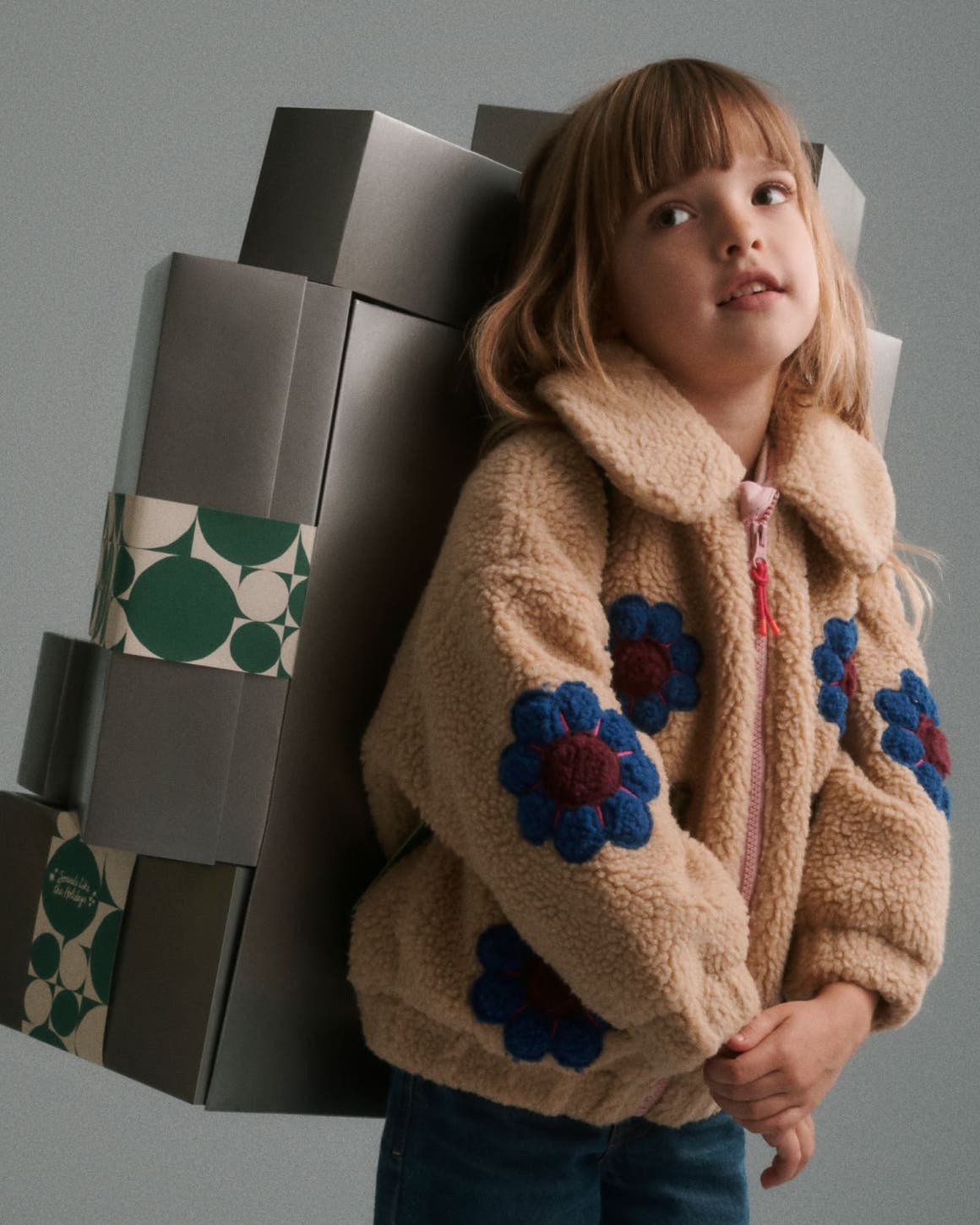 A girl in a fleece jacket carrying a stack of silver Nordstrom gift boxes on her back.