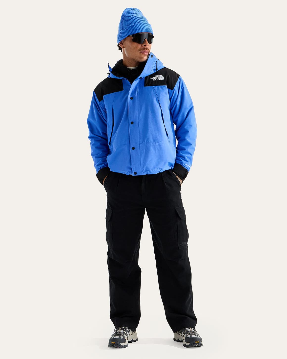 A man in a blue The North Face jacket, black cargo pants, sunglasses and hiking sneakers.