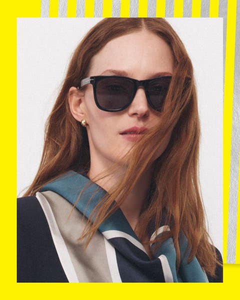A woman wearing sunglasses, a navy suit & draped scarf.