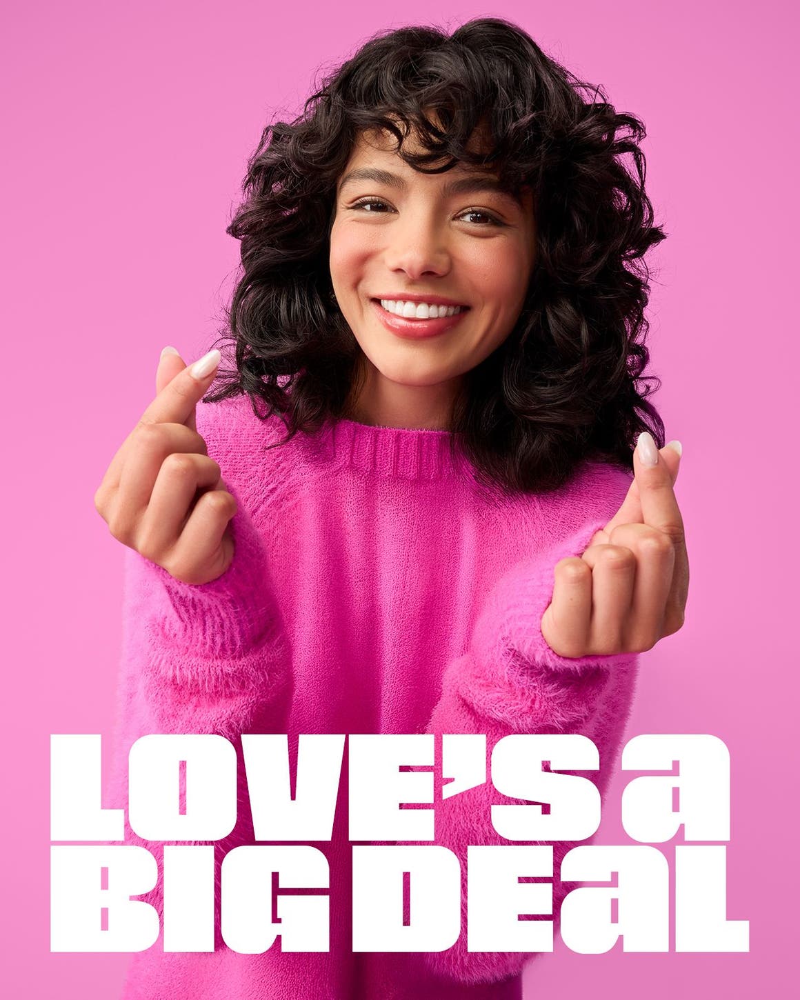 A woman in a pink crewneck sweater holds up both hands, making heart shapes with her thumb and pointer fingers. 