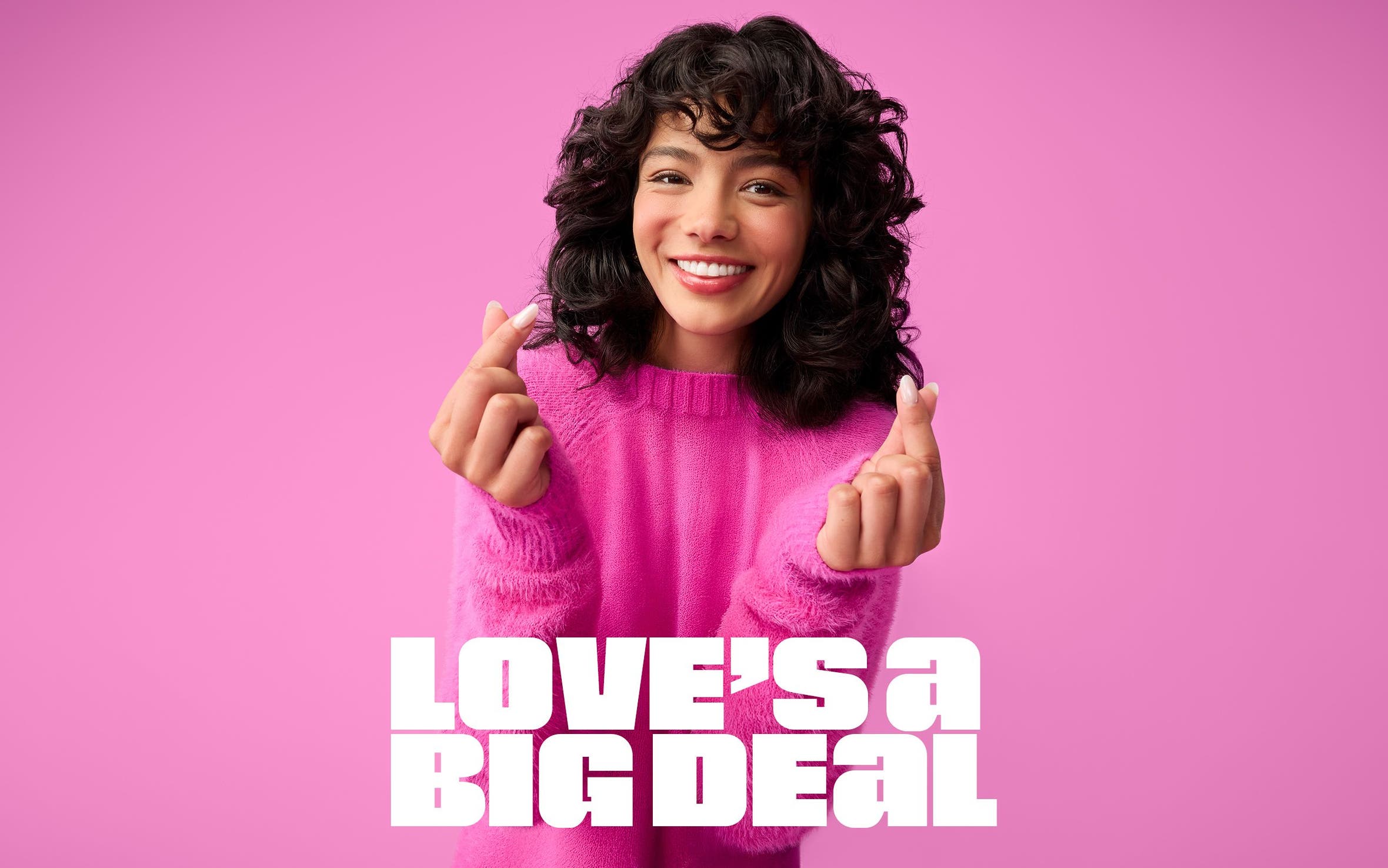 A woman in a pink crewneck sweater holds up both hands, making heart shapes with her thumb and pointer fingers. 
