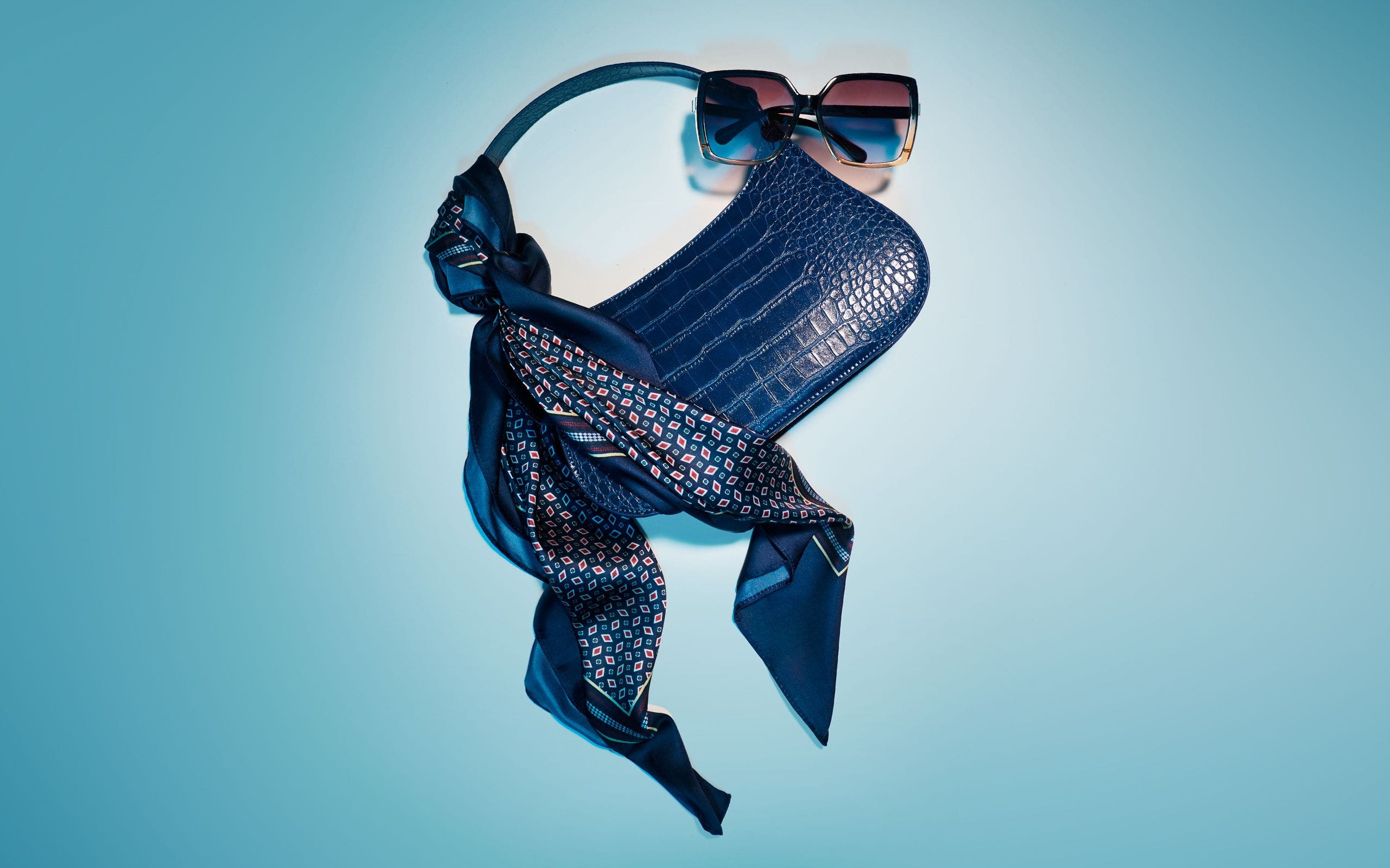 A dark blue croc-embossed shoulder bag styled with a printed scarf and sunglasses.