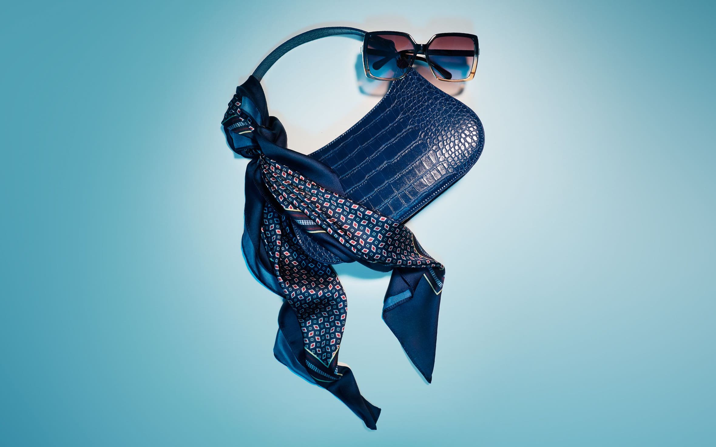A dark blue croc-embossed shoulder bag styled with a printed scarf and sunglasses.