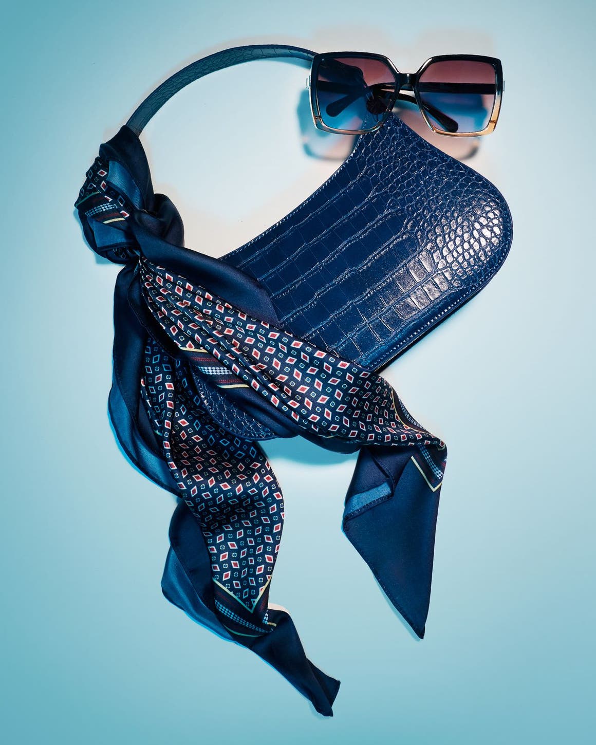 A dark blue croc-embossed shoulder bag styled with a printed scarf and sunglasses.