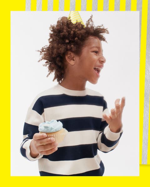 A boy wearing a striped sweater and party hat with a cupcake.
