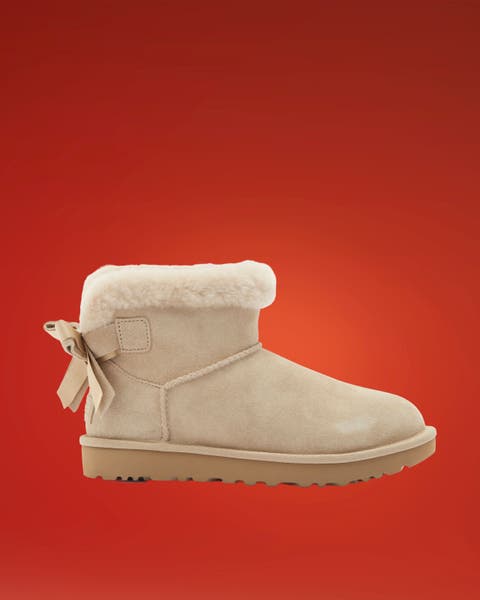 Sand suede UGG boot with shearling cuff and back bow on red.