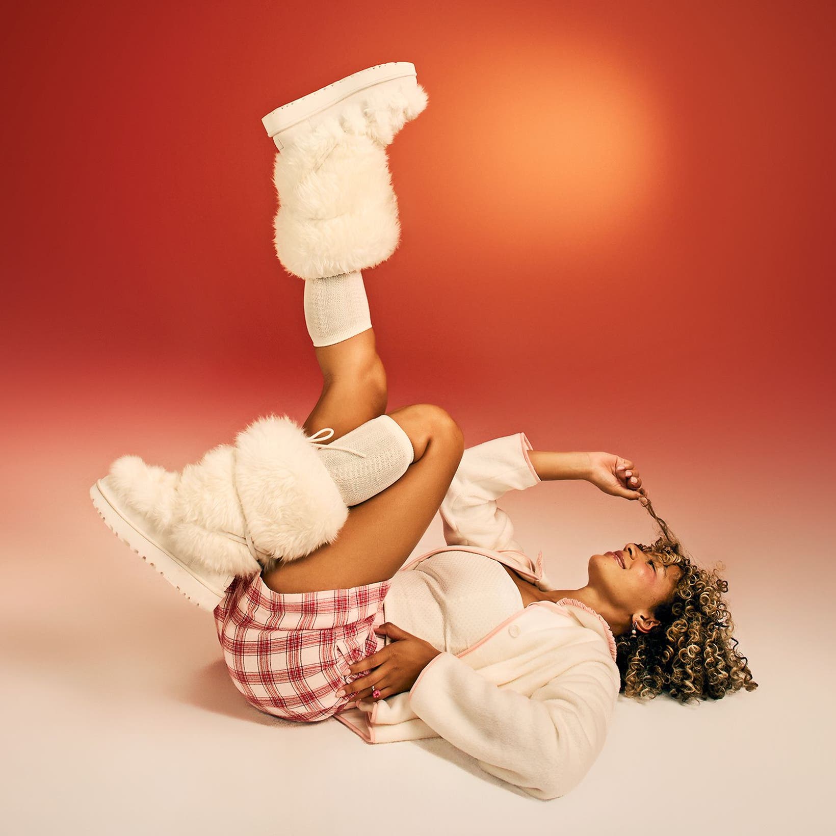 A woman wearing fuzzy boots and cozy clothing poses on the floor with her feet in the air.
