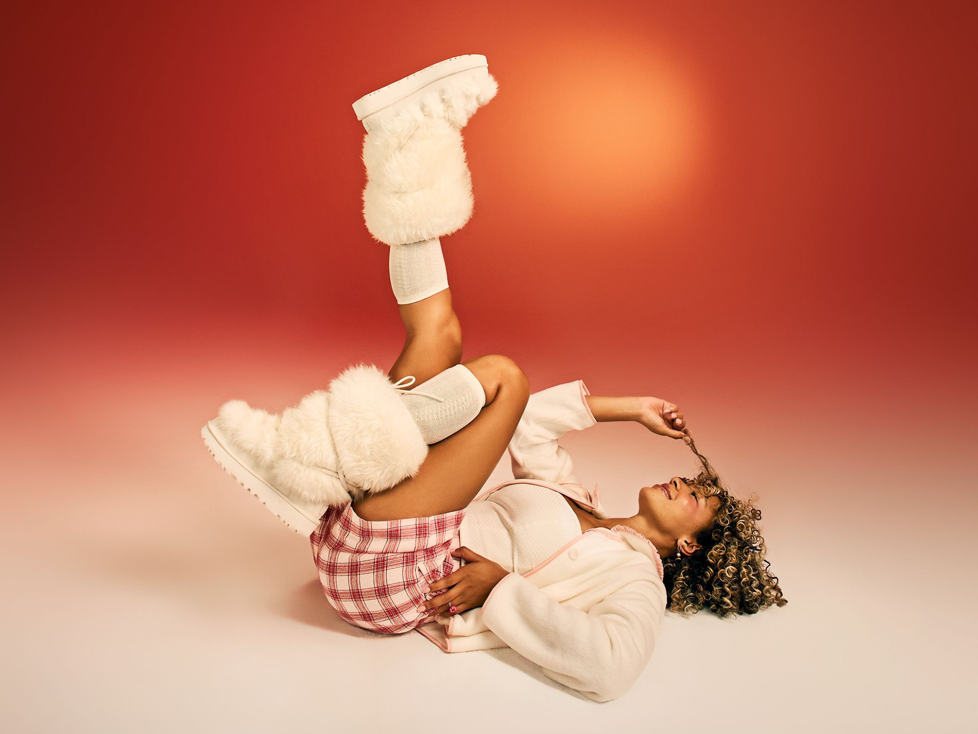 A woman wearing fuzzy boots and cozy clothing poses on the floor with her feet in the air.