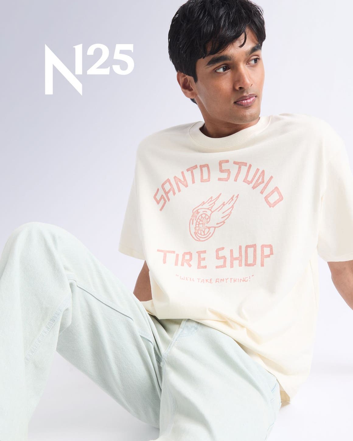 A man sitting wearing Santo Studio tee, jeans, and sneakers.