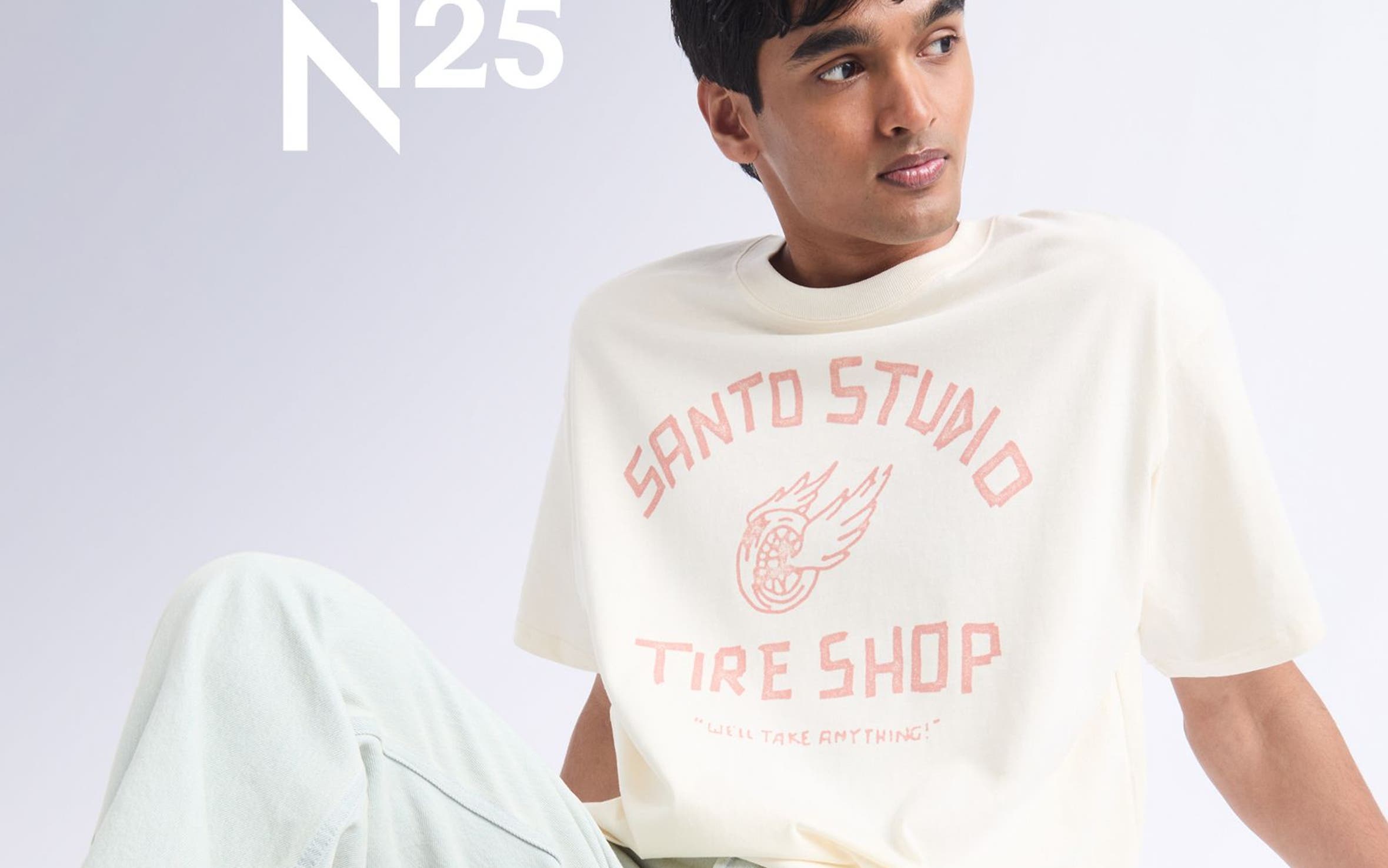 A man sitting wearing Santo Studio tee, jeans, and sneakers.