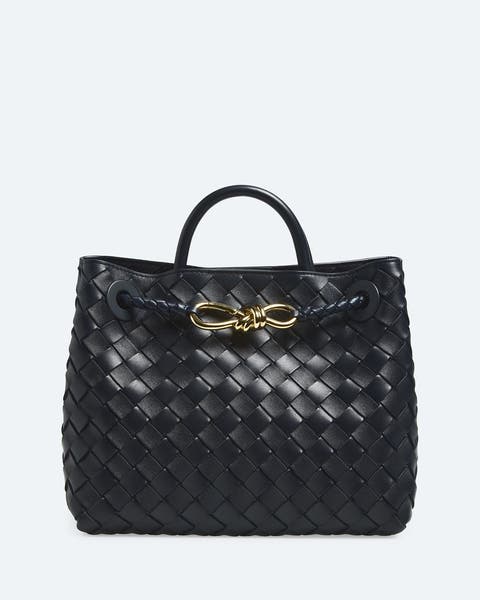 Black woven leather tote bag with a top handle and a gold-tone knot-shaped hardware accent at the front.