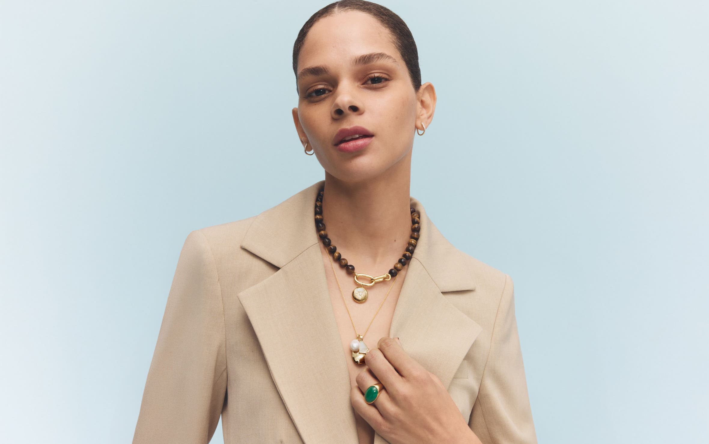 A woman in a linen blazer wearing a gold charm necklace, a wooden bead choker and a gold and green stone statement ring.