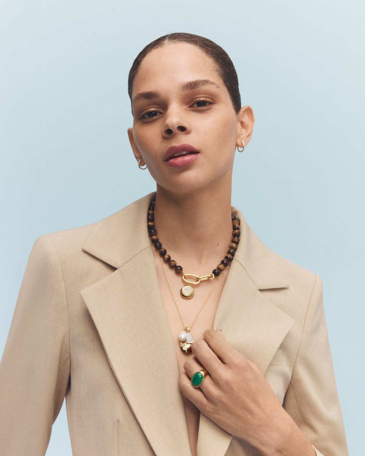 A woman in a linen blazer wearing a gold charm necklace, a wooden bead choker and a gold and green stone statement ring.