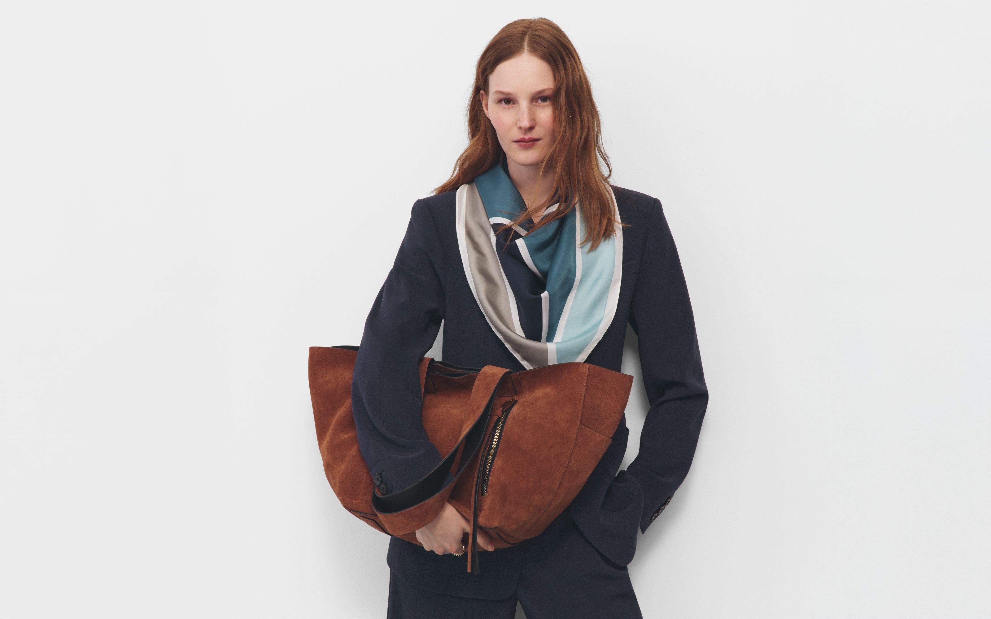 A woman wearing a navy suit, draped scarf and holding a large half-moon caramel suede bag.