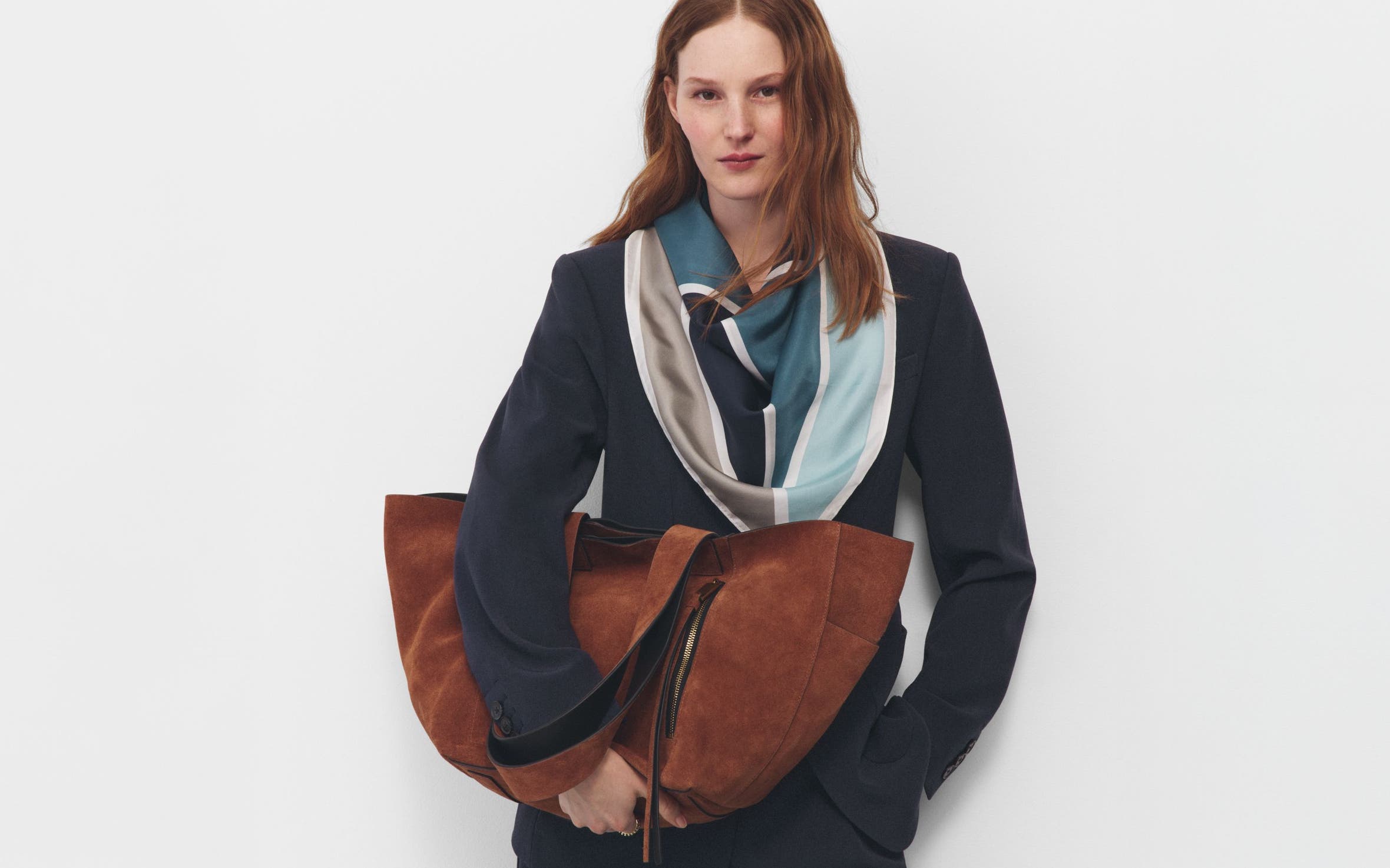 A woman wearing a navy suit, draped scarf and holding a large half-moon caramel suede bag.
