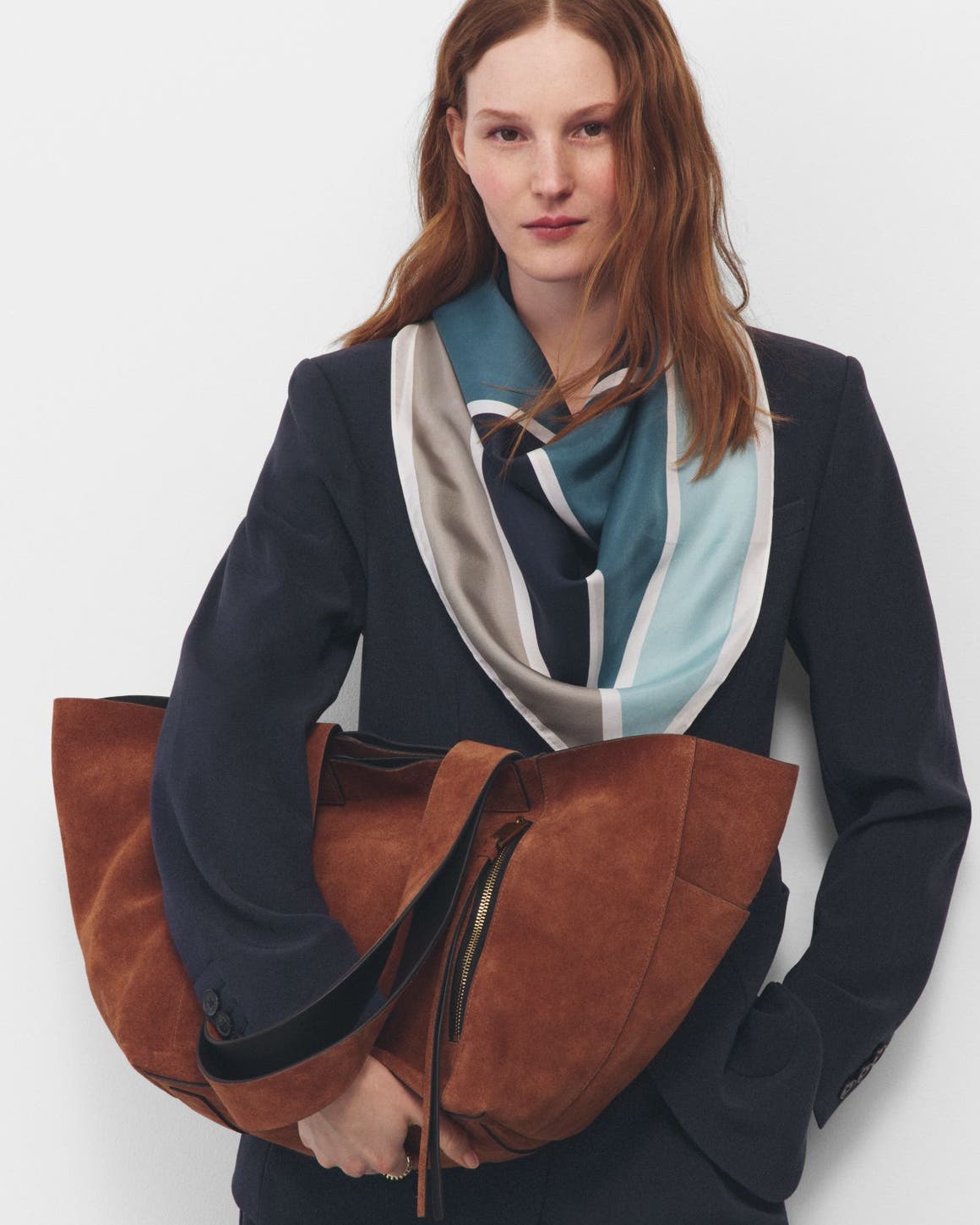 A woman wearing a navy suit, draped scarf and holding a large half-moon caramel suede bag.