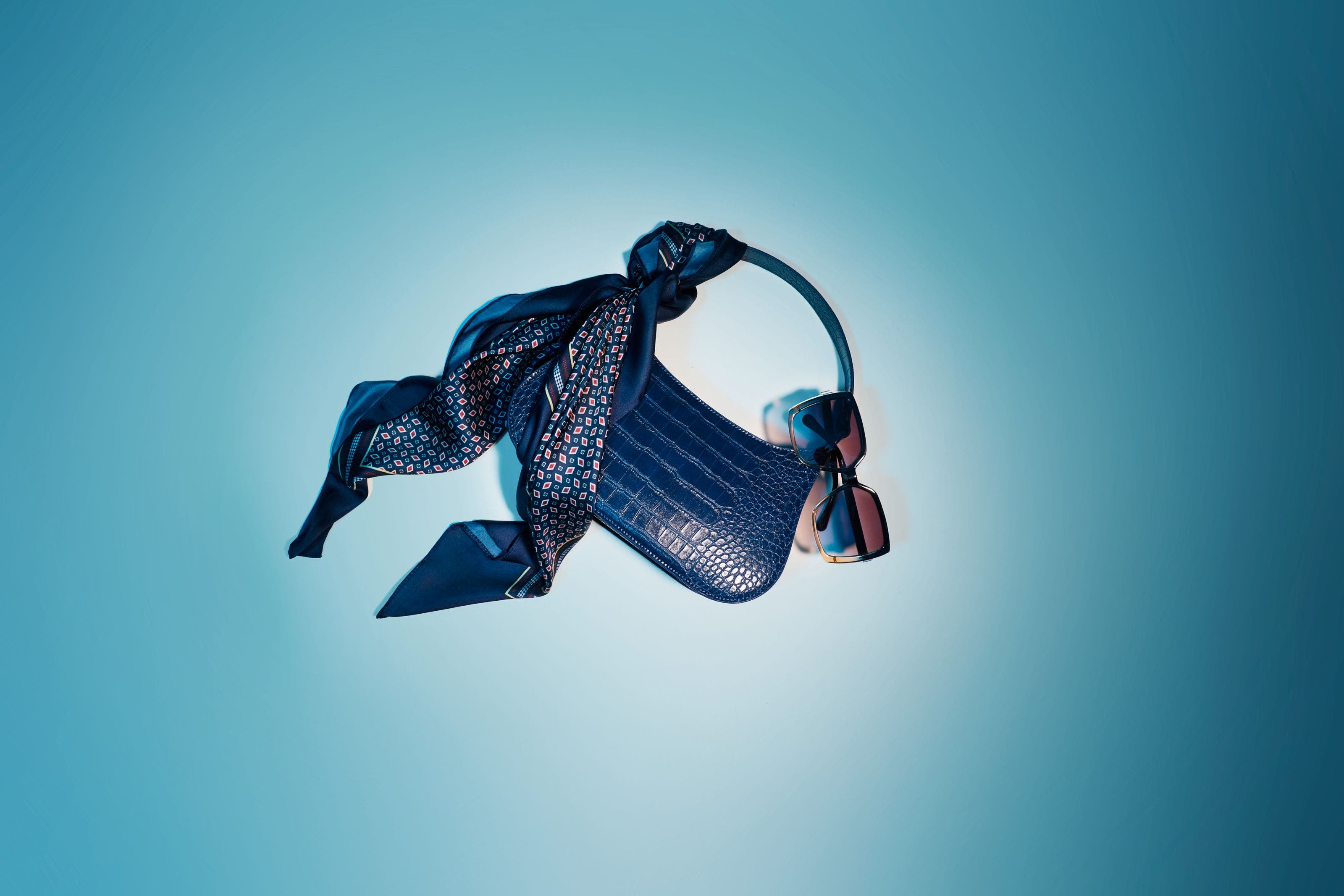 Blue croc-embossed handbag with a navy patterned scarf tied to the handle, paired with pink-lensed sunglasses.