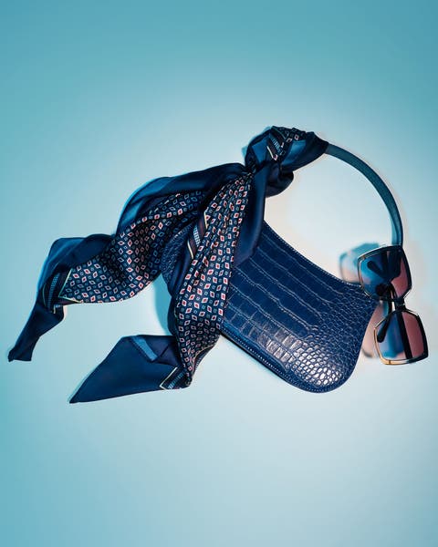 Blue croc-embossed handbag with a navy patterned scarf tied to the handle, paired with pink-lensed sunglasses.