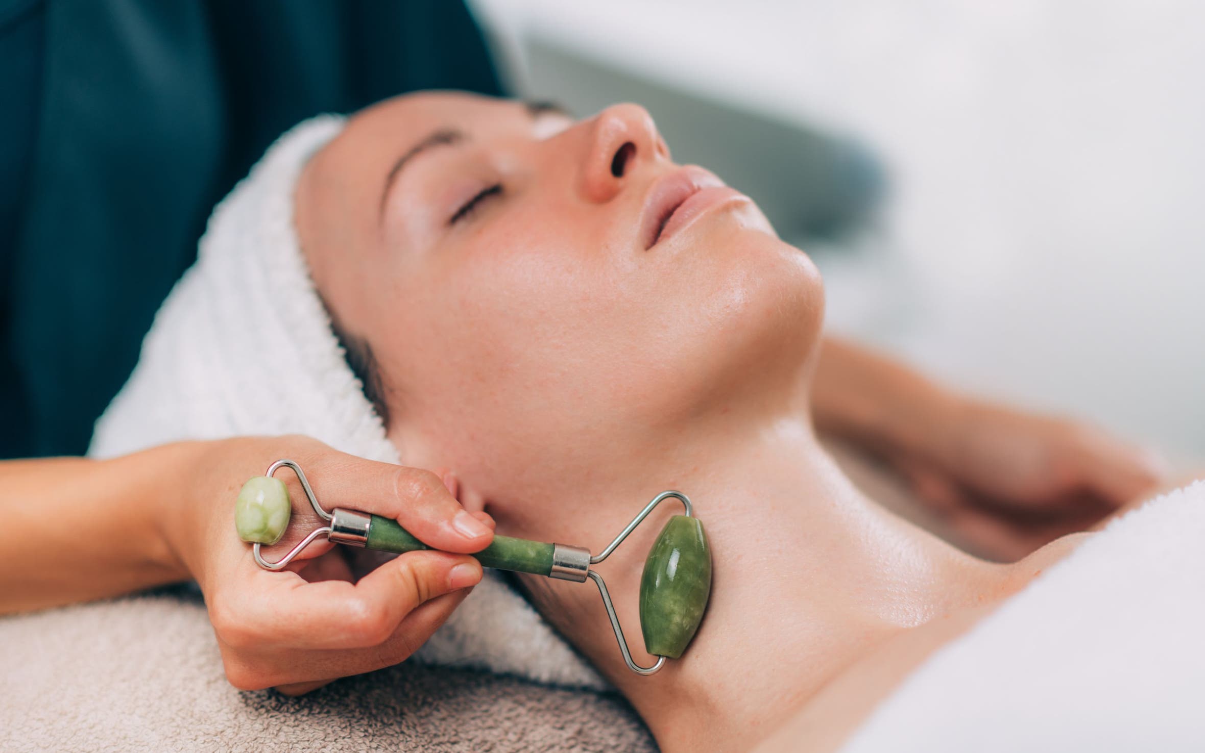 A woman with her eyes closed and her hair wrapped in a towel, getting a jade roller facial massage along her neck.