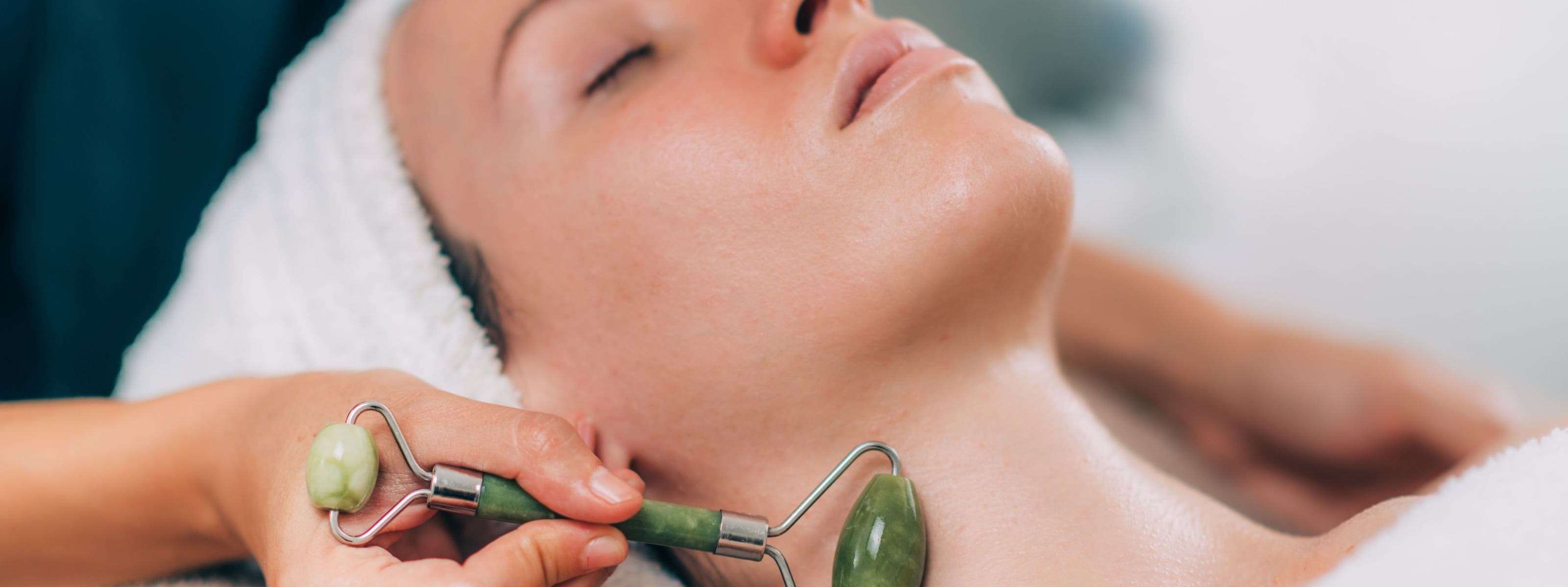 A woman with her eyes closed and her hair wrapped in a towel, getting a jade roller facial massage along her neck.