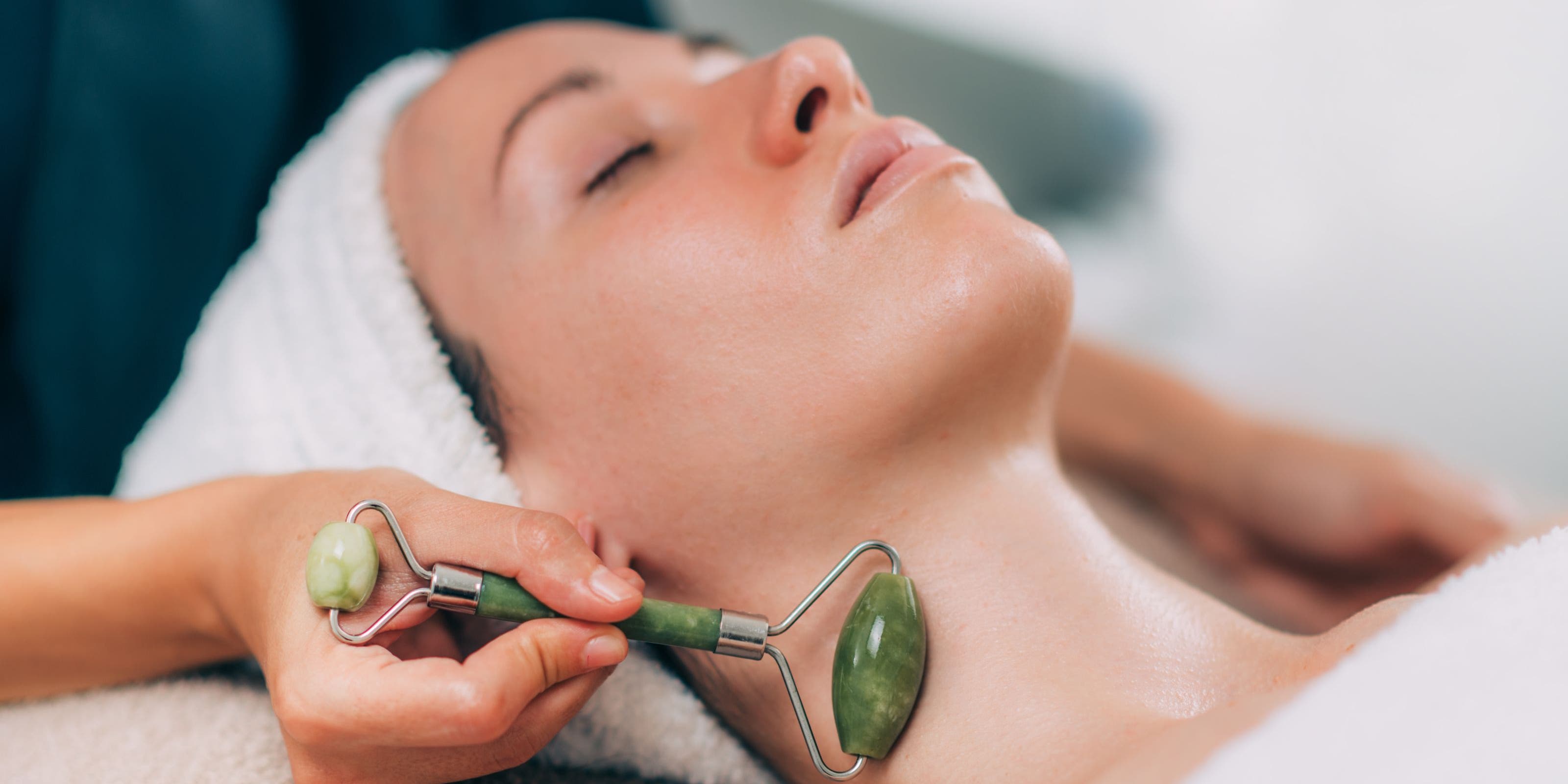 A woman with her eyes closed and her hair wrapped in a towel, getting a jade roller facial massage along her neck.