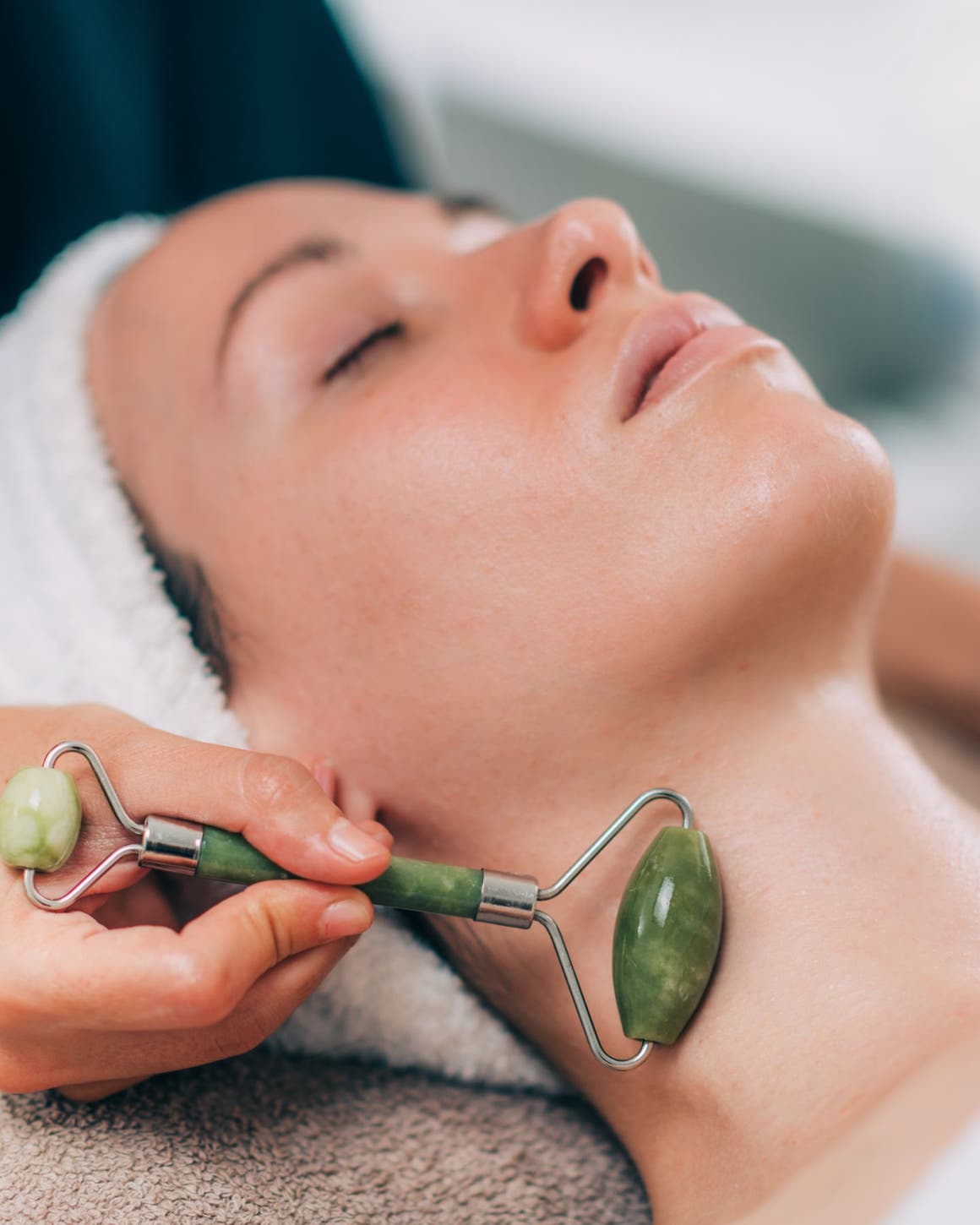 A woman with her eyes closed and her hair wrapped in a towel, getting a jade roller facial massage along her neck.