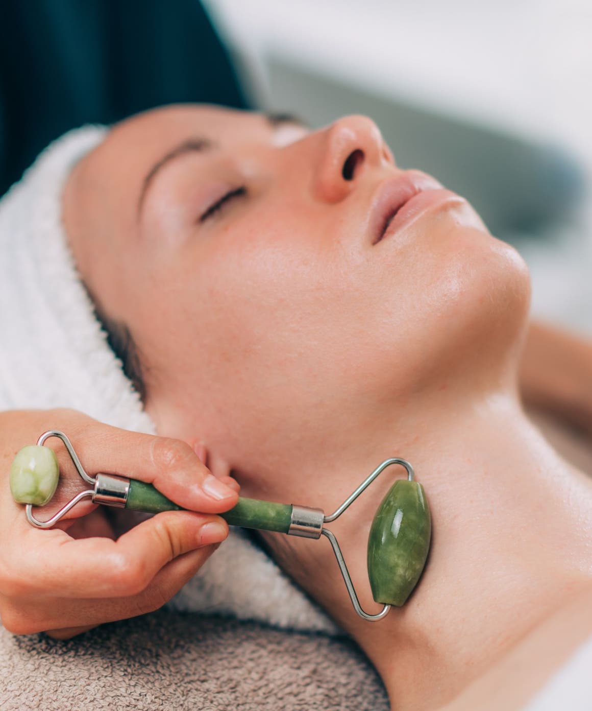 A woman with her eyes closed and her hair wrapped in a towel, getting a jade roller facial massage along her neck.